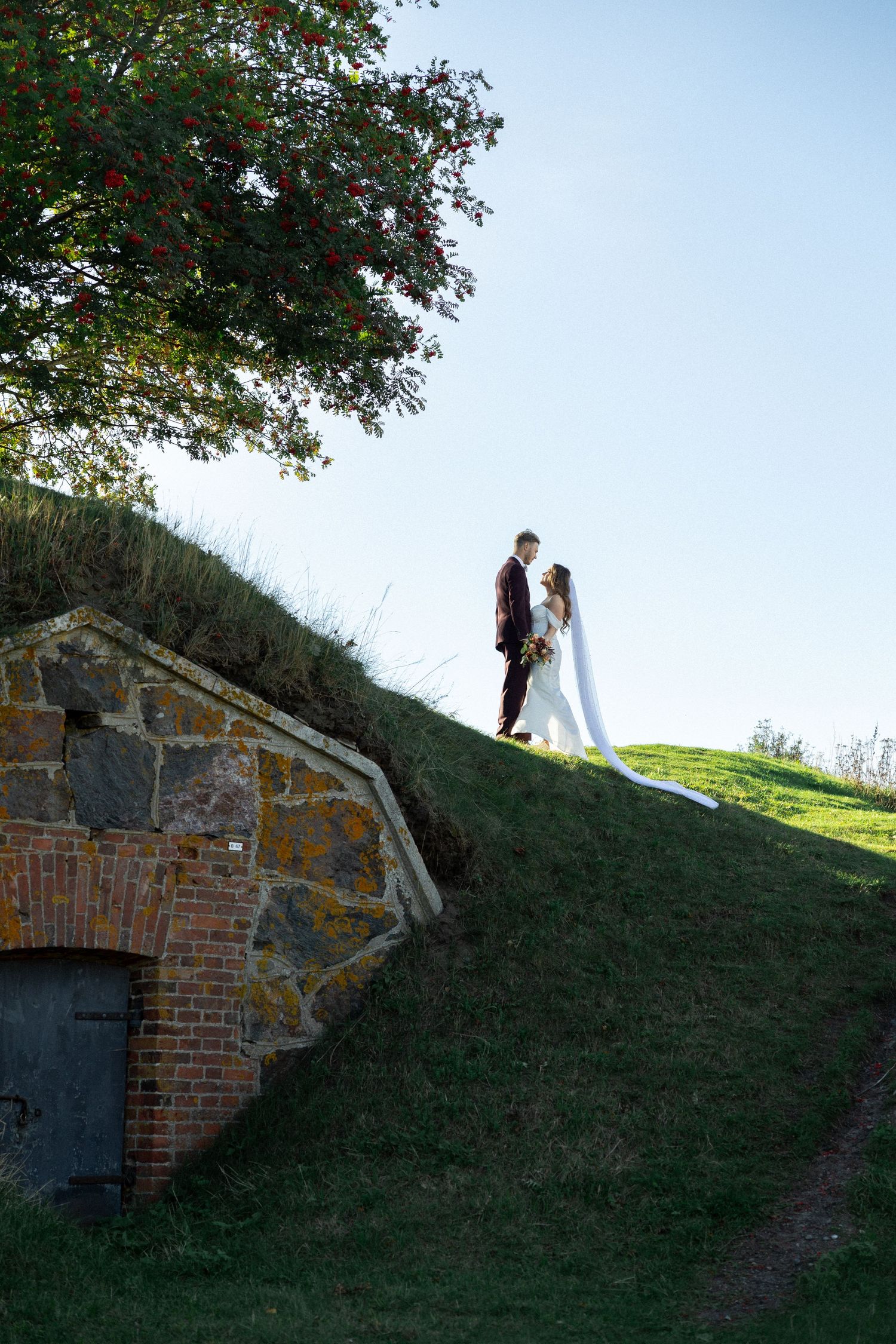 A wedding couple standing on a grassy hill above a stone archway.