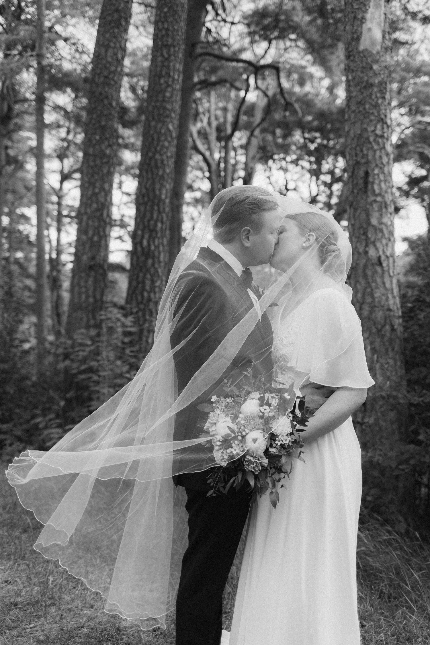 Black and white portrait of newlyweds with flowing veil against forest backdrop.