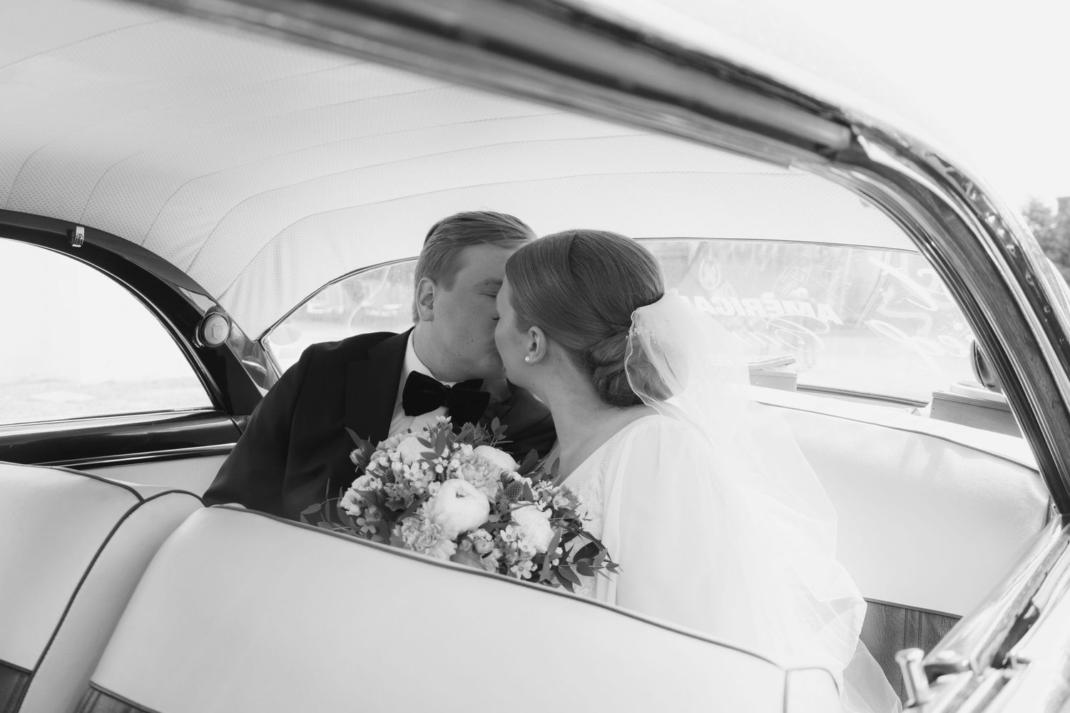 Intimate wedding moment captured through vintage car window with floral bouquet visible.