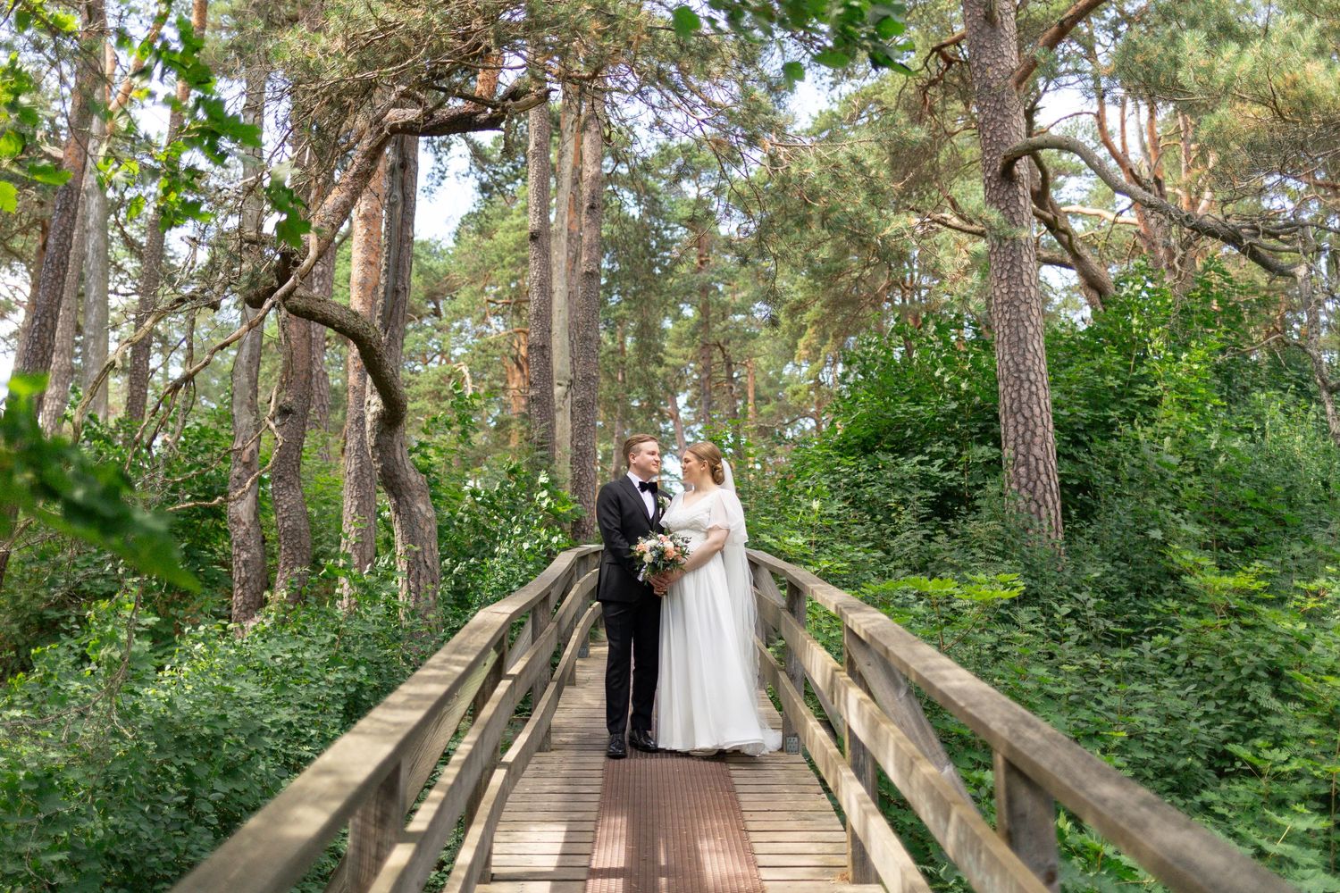 Newlywed couple poses on wooden boardwalk surrounded by tall pine trees.