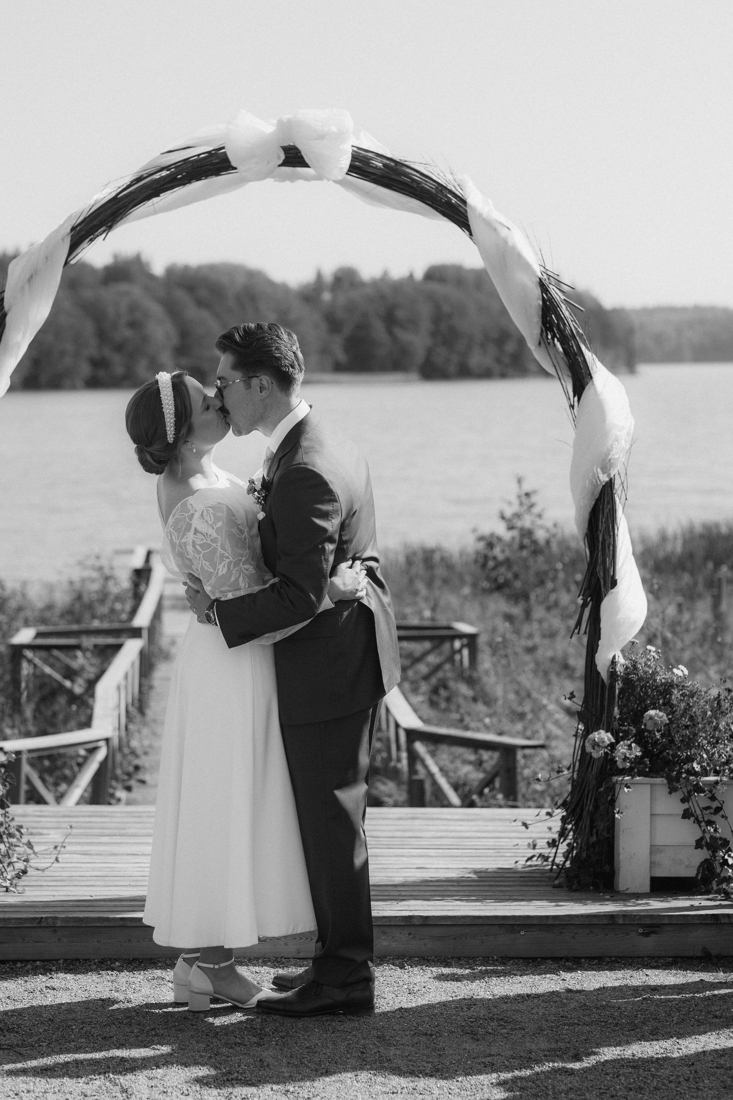 Black and white photo of a couple embracing under a decorated wedding arch by a lake.