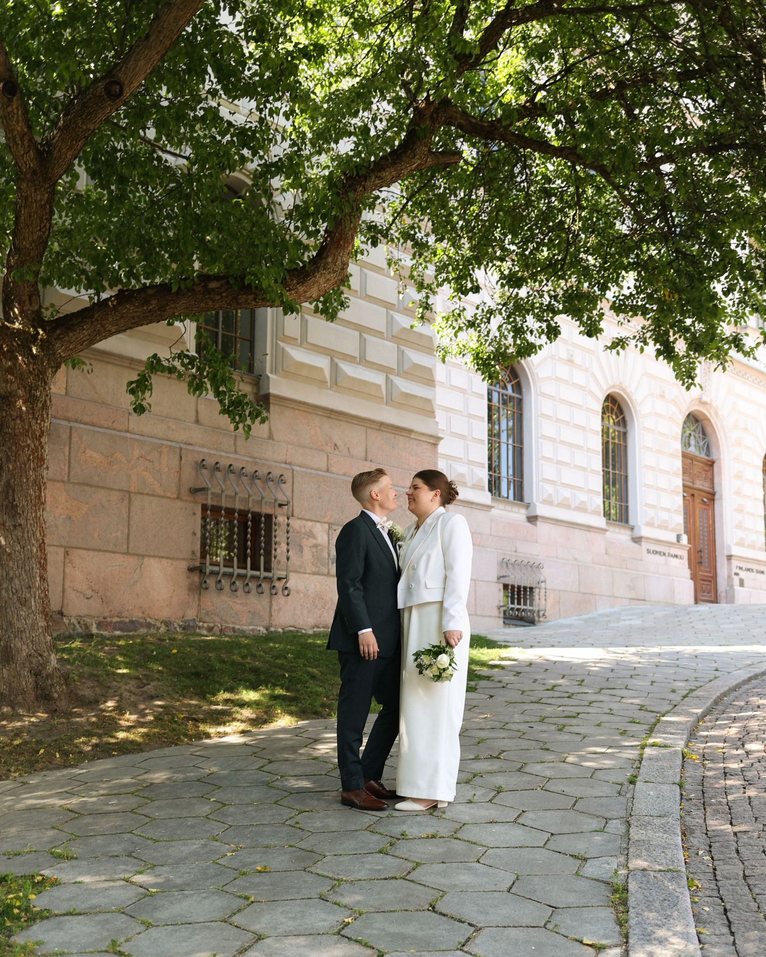 Romantic wedding portrait under green tree branches next to historic building.