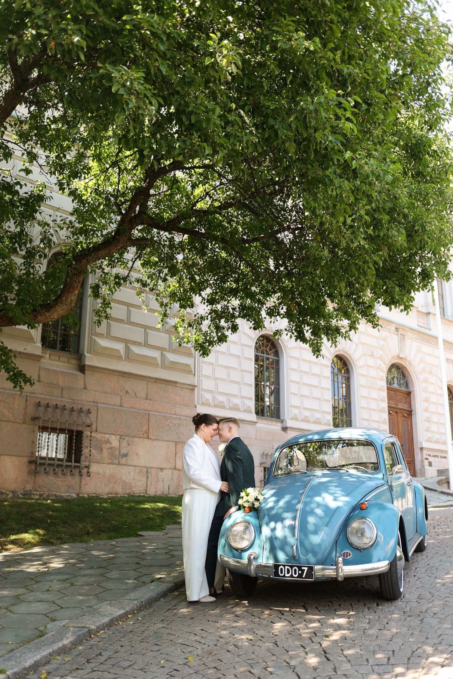 Wedding couple posing by vintage blue Volkswagen Beetle near historic building.
