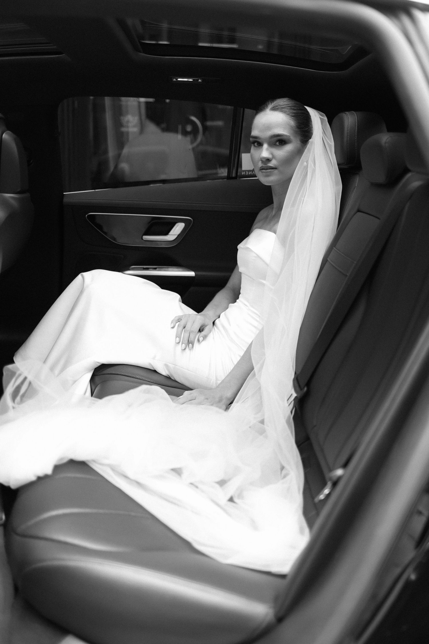 A bride in a white gown sits elegantly in the backseat of a luxury car.