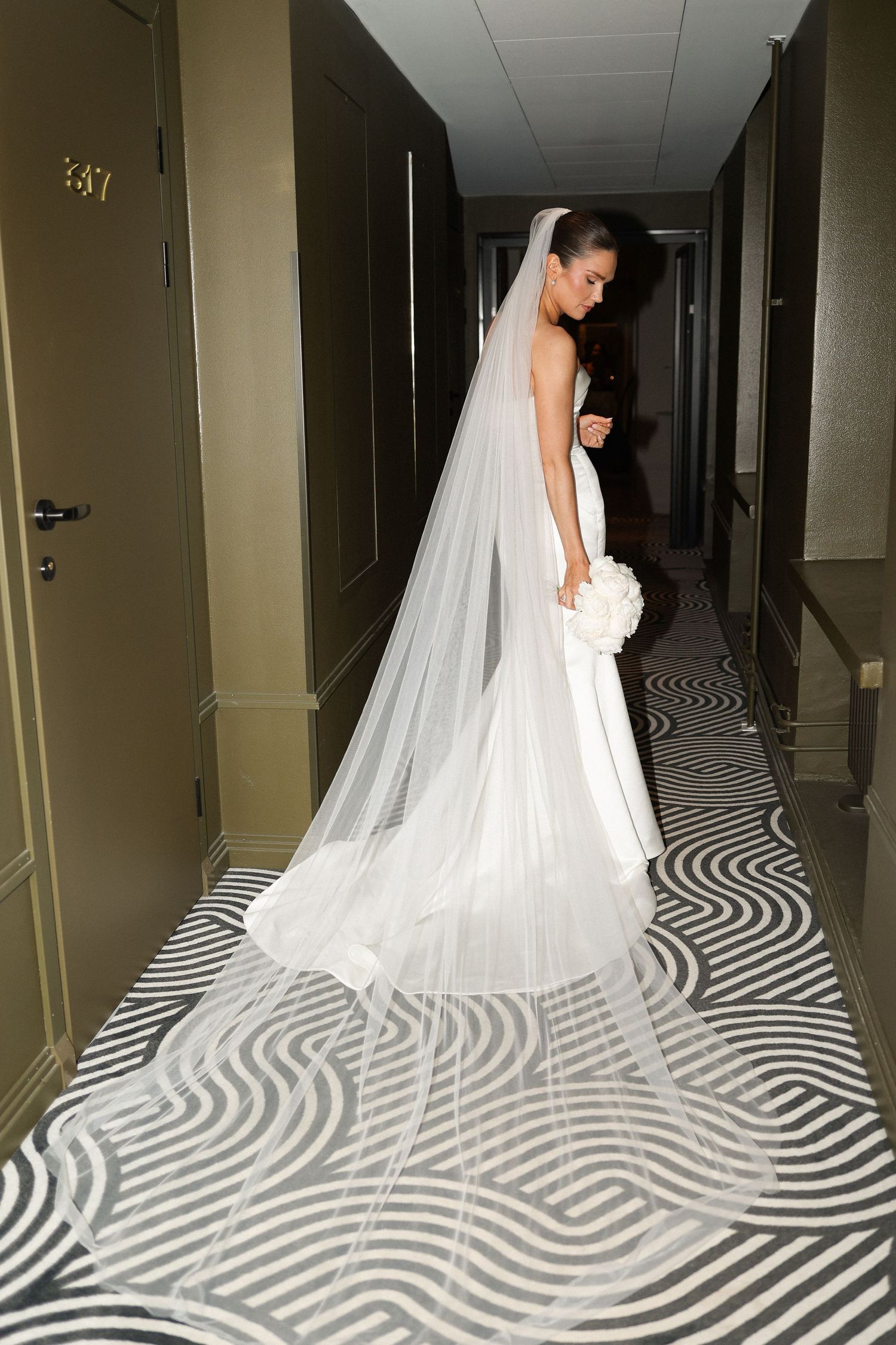 A bride in a flowing cathedral veil poses in a modern hotel hallway.
