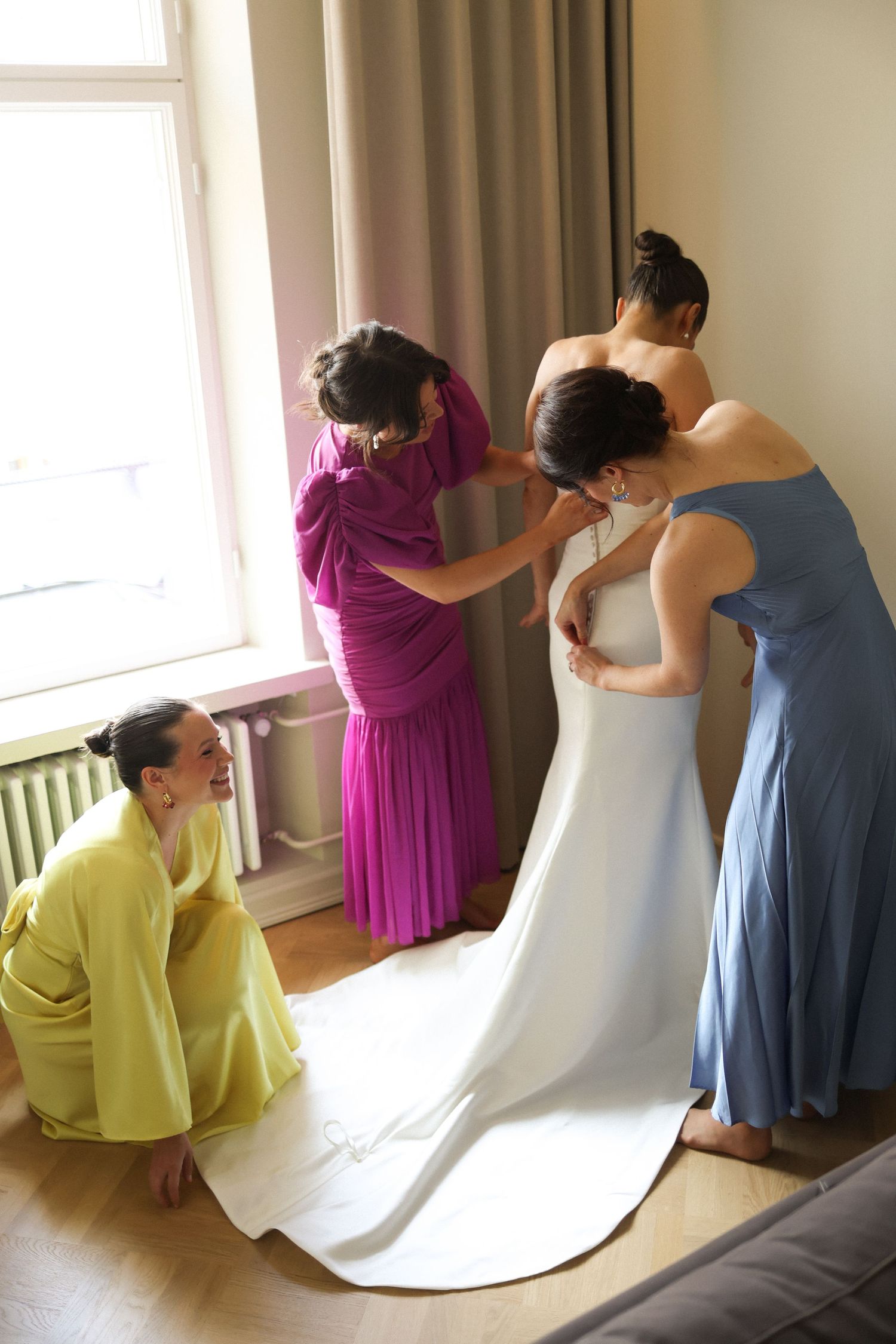 Bridesmaids in colorful dresses help the bride get ready by a sunny window.