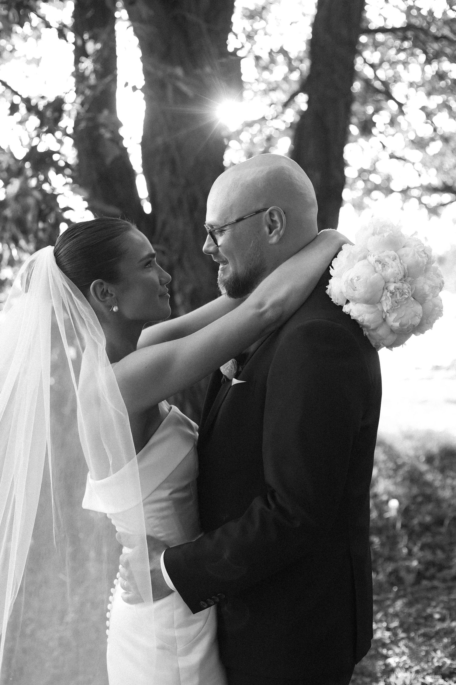 Romantic black and white portrait of newlyweds embracing under trees with sunlight streaming through.