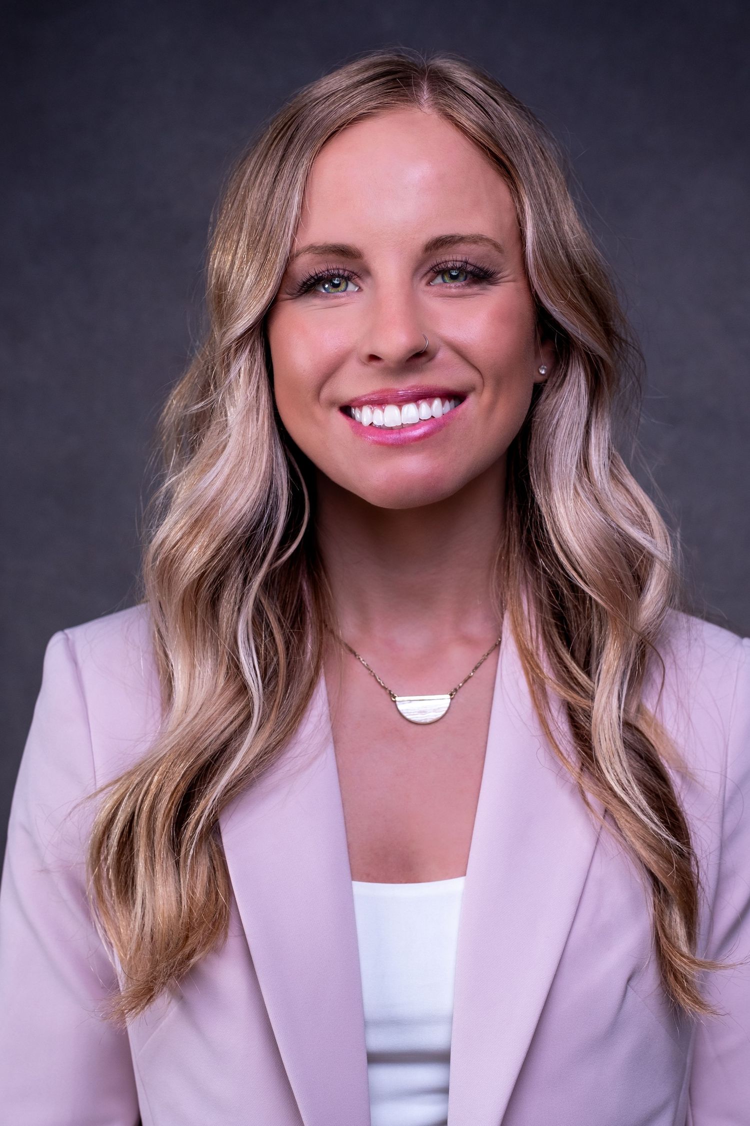 Professional headshot of someone with wavy blonde hair wearing a blush pink blazer and white top with a delicate necklace.