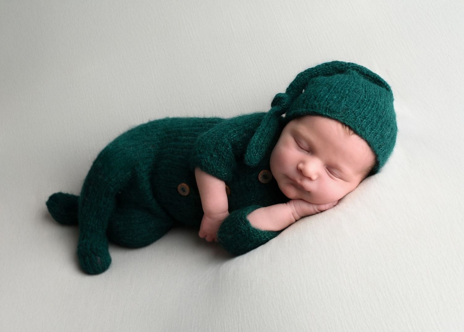 Sleeping newborn wearing emerald green knitted outfit and hat on white background.