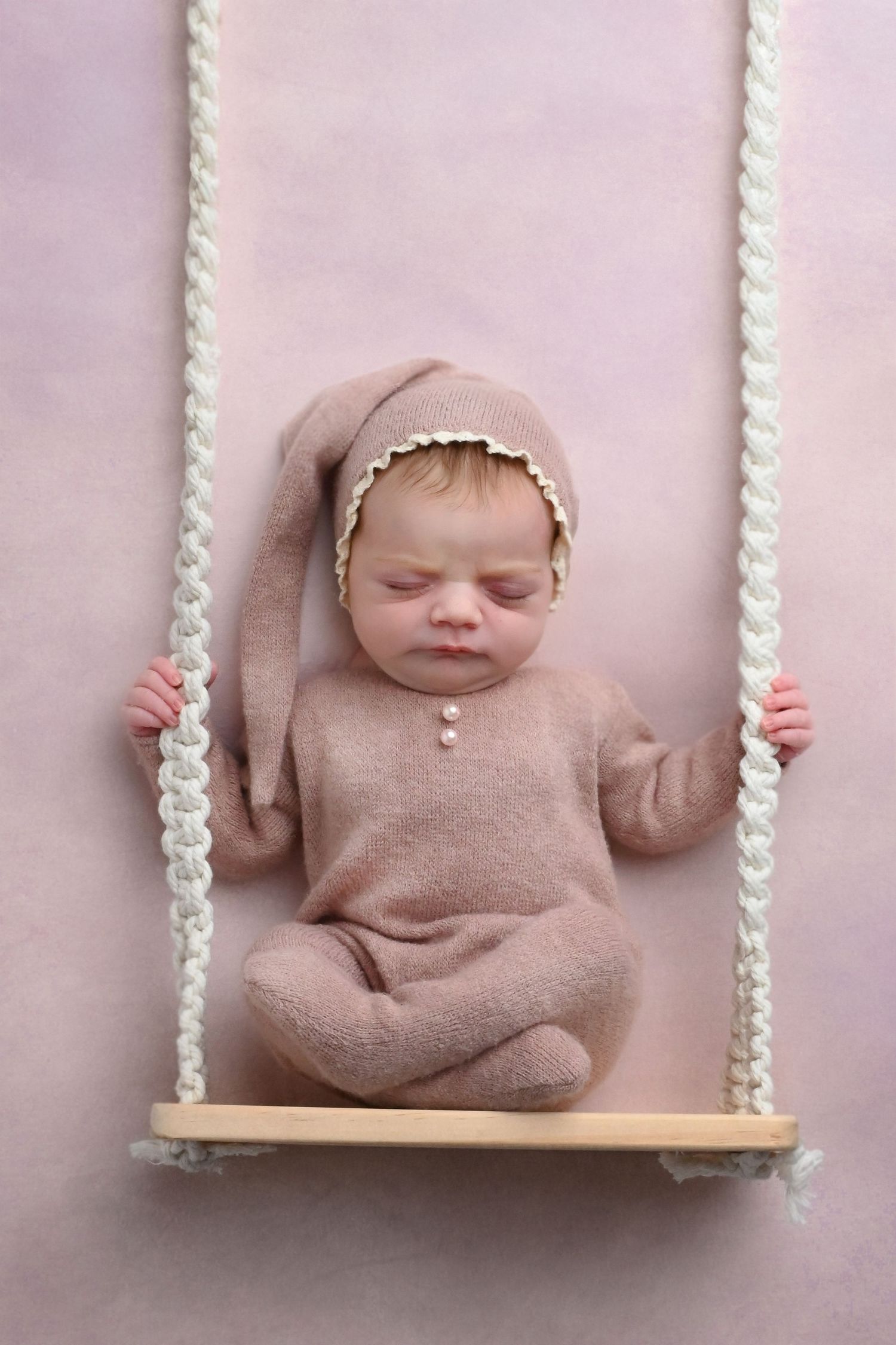 Baby in dusty pink outfit posed on decorative rope swing against pink backdrop.