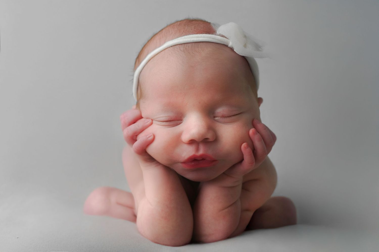 Artistic newborn photography with baby posed on minimalist gray background with white headband.