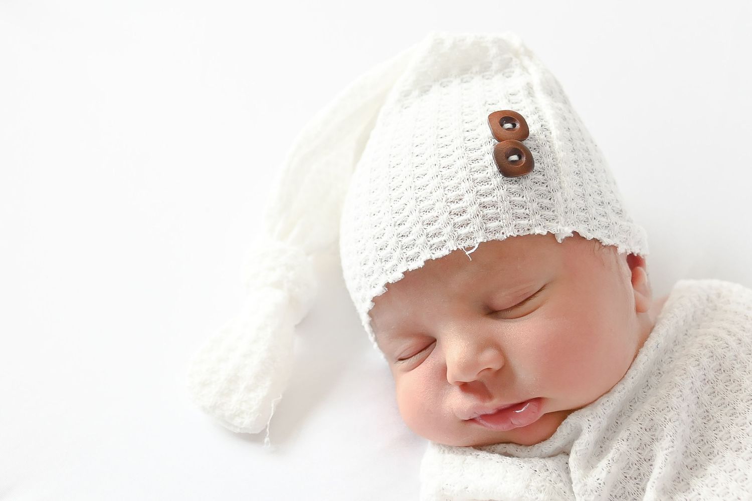 Sleeping newborn baby wearing soft white knit bonnet captured in gentle lighting.
