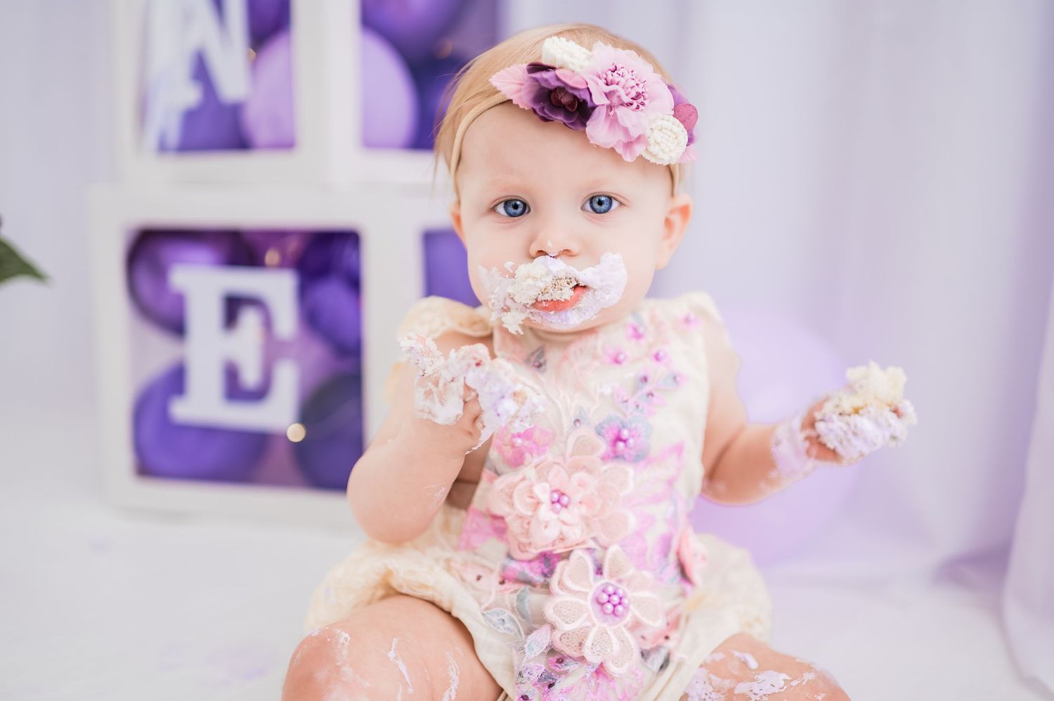 Baby in floral outfit sits among purple decorations during a milestone photoshoot session.