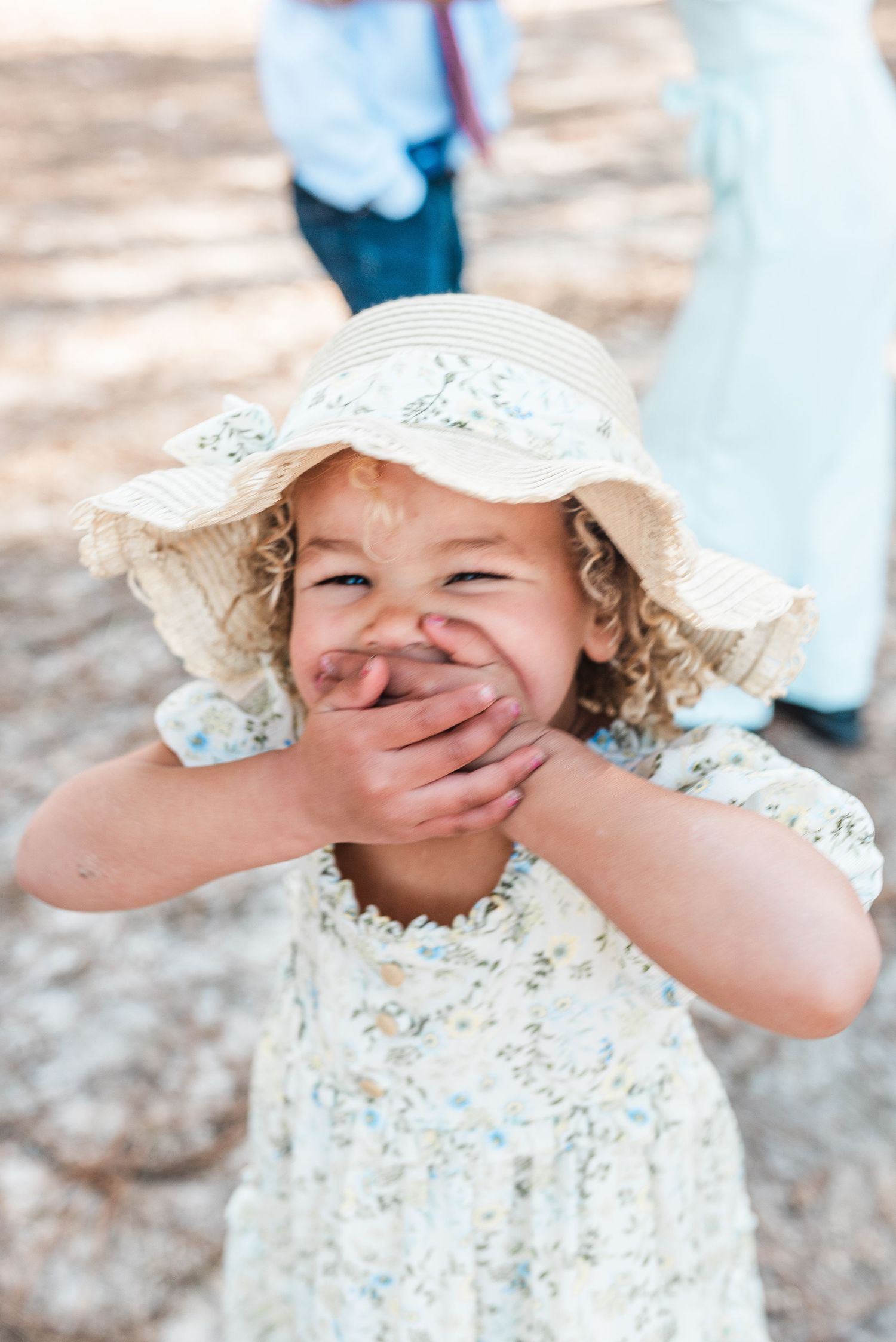 A child in a floral dress and sun hat covers their mouth while giggling.
