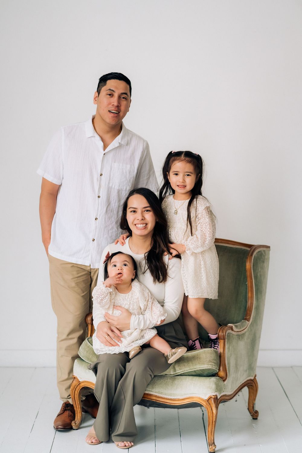 Family wearing coordinated white outfits poses together around a vintage green armchair in a bright studio setting.