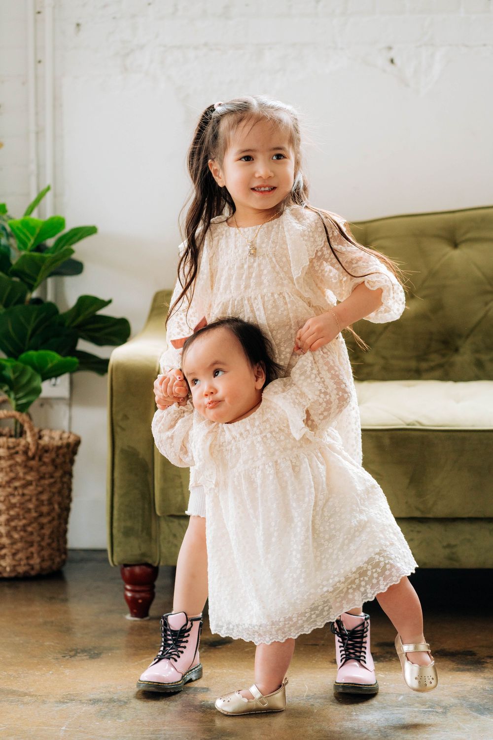 Two children in matching white lace dresses with contrasting footwear stand together in a cozy living room interior.