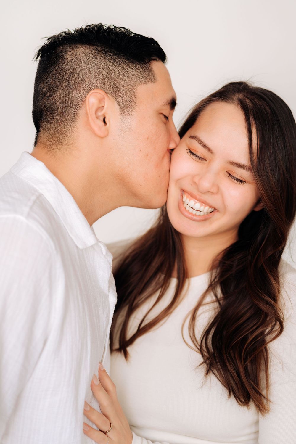 A loving couple wearing white outfits share a tender moment as one kisses the other's cheek while she smiles joyfully.