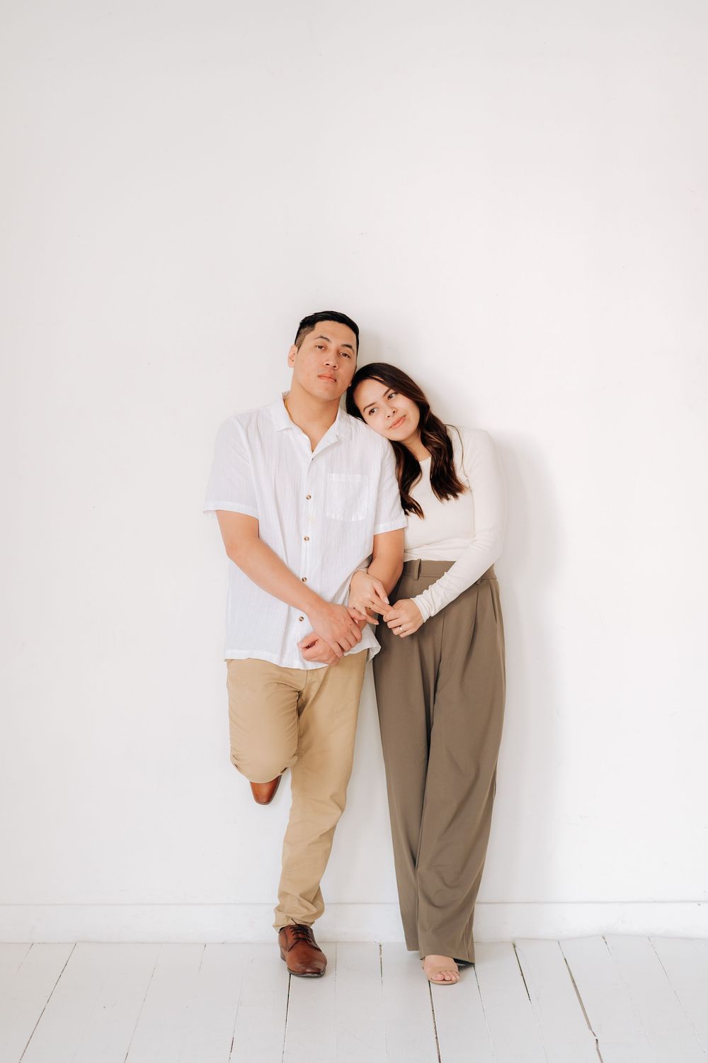 A couple leans on a white wall wearing coordinated outfits of white shirt and khaki pants, creating a minimalist feel.