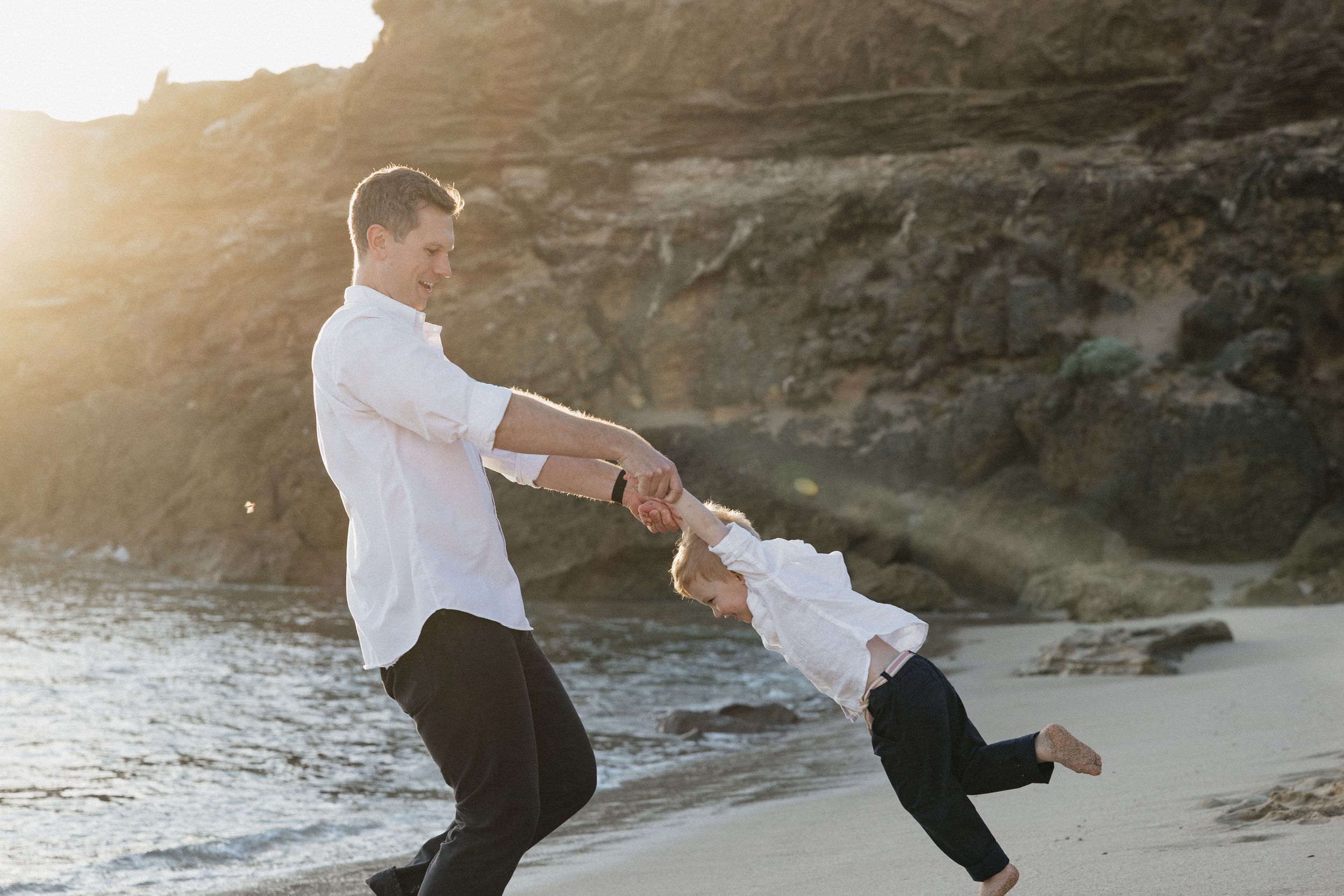 A father joyfully swings his child by the hands on a sunny beach with rocky cliffs in the background.