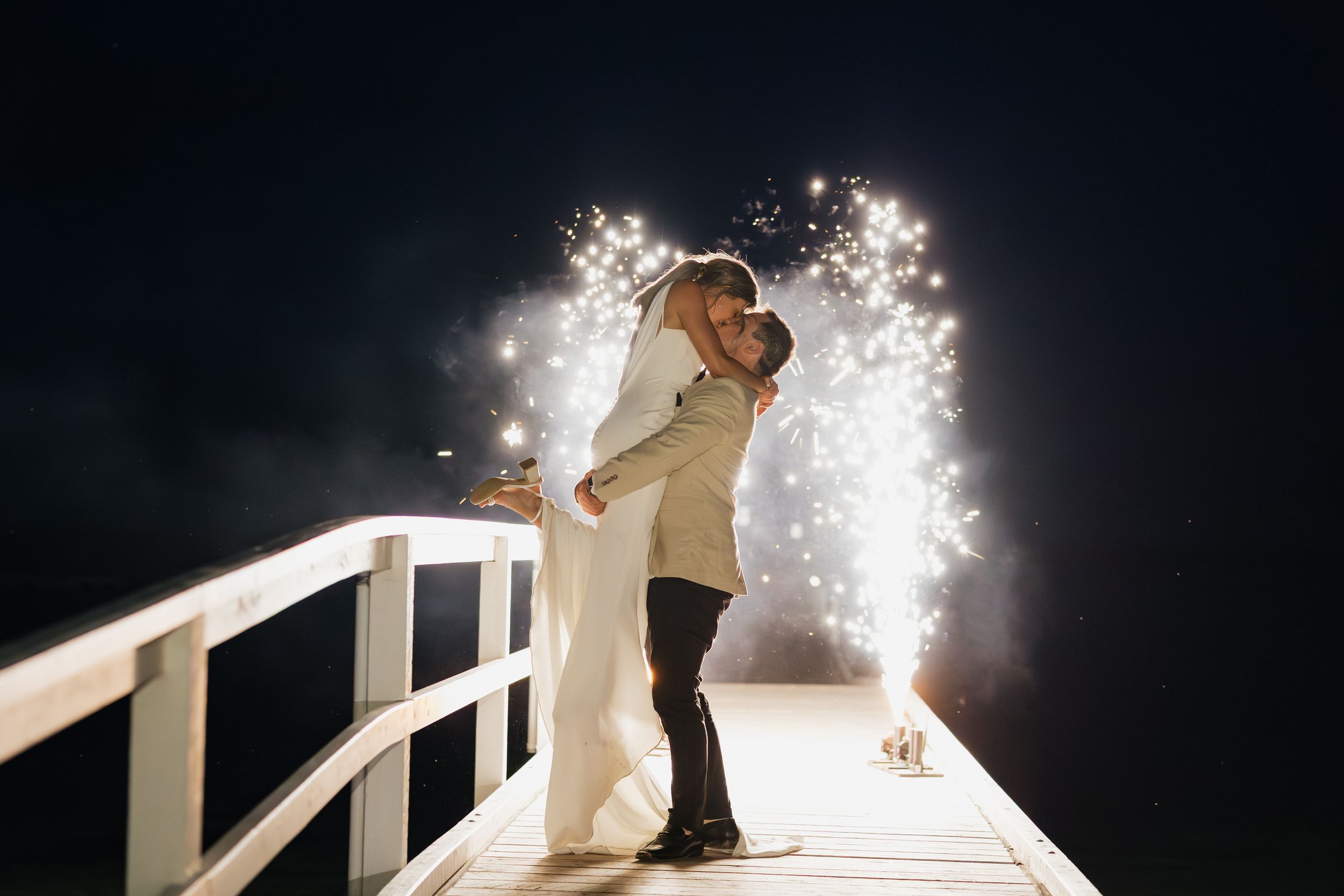 Bride and groom kissing on a pier with sparkling fireworks at night during their romantic wedding celebration.