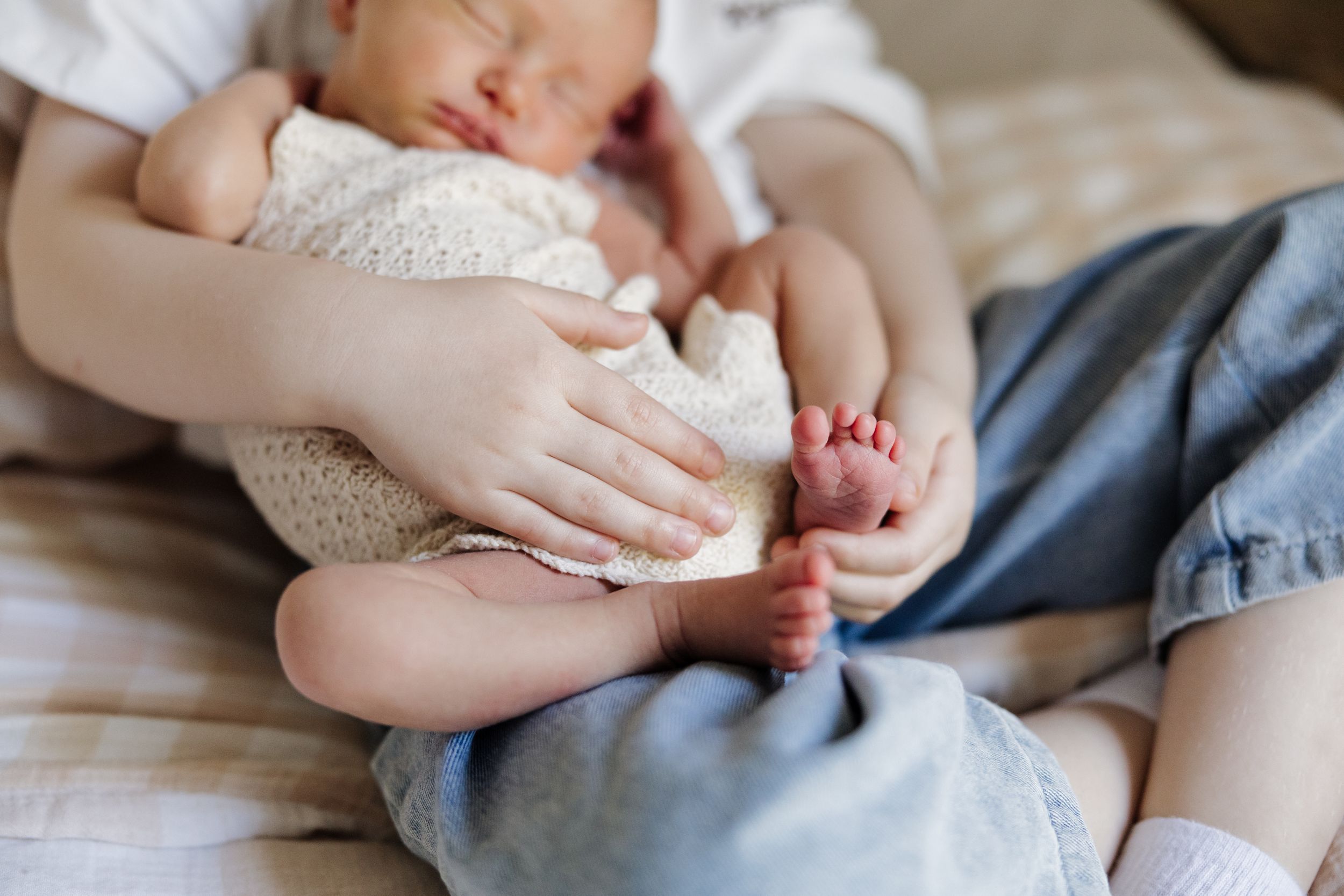 Older child gently holding a sleeping newborn baby wrapped in a white knit blanket, showing tiny feet.