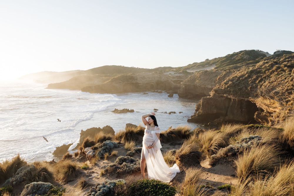 A woman in a flowing white dress stands on coastal dunes with golden sunlight illuminating a dramatic ocean cliff backdrop.