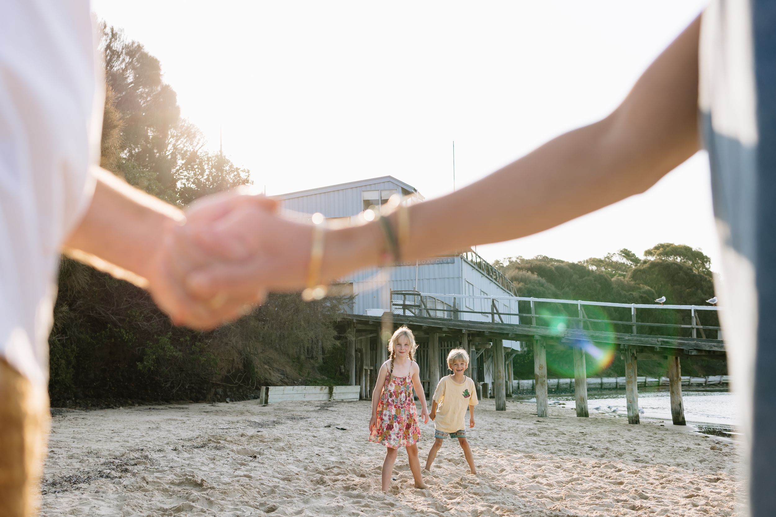 Two children walk on a sandy beach as parents hold hands in the foreground with a modern building behind.