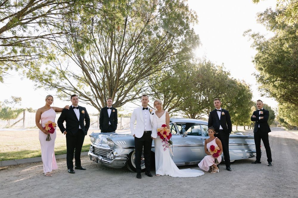 Wedding party posing with a vintage silver car outdoors, bride and groom with bridal party in formal attire under trees.