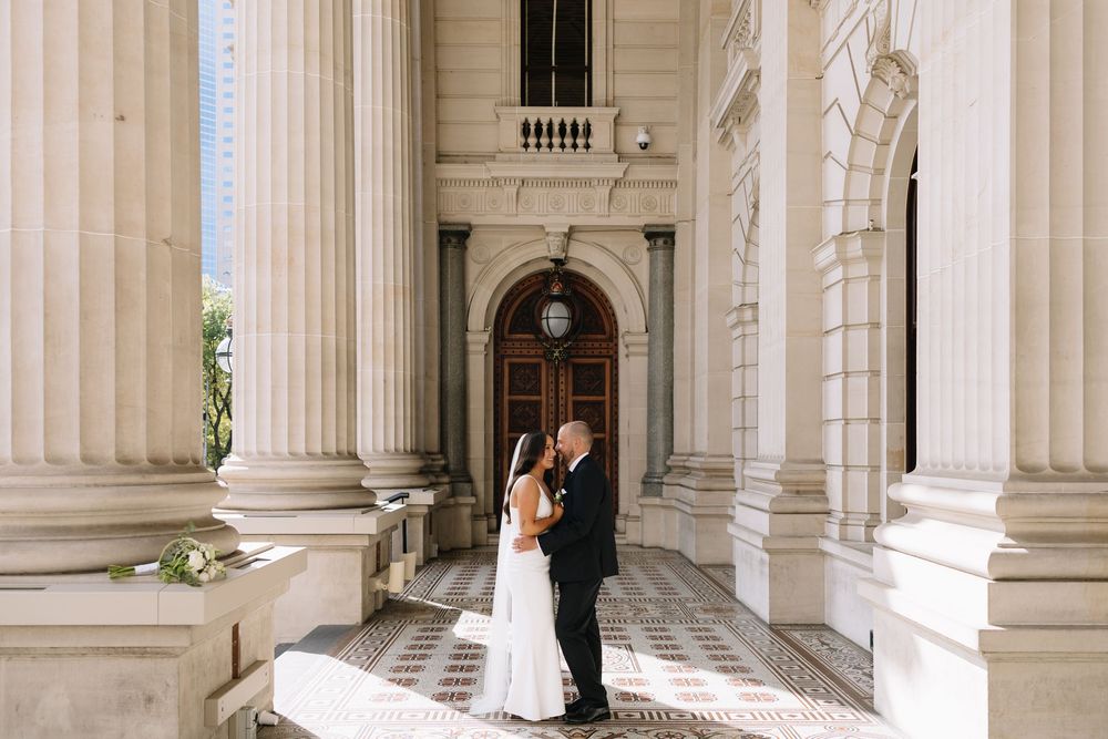 Bride and groom share a romantic moment among grand white columns at an elegant neoclassical building wedding venue.
