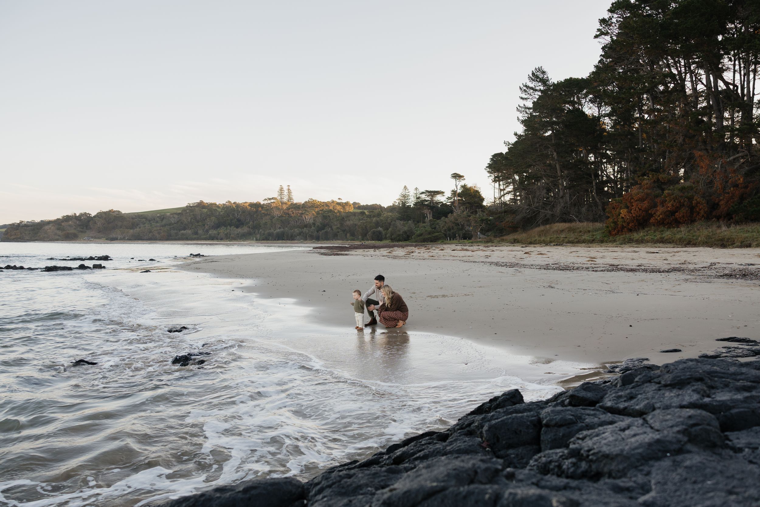 Couple sitting together on a sandy beach shore with rocky coastline and tall pine trees in the background.