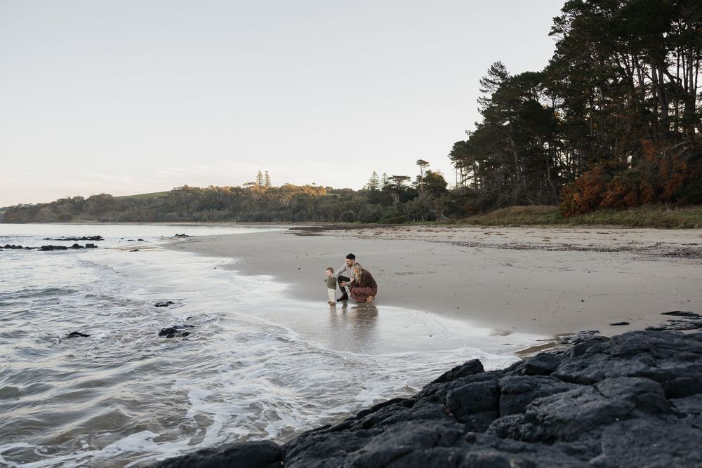 Couple sitting together on a sandy beach shore with rocky coastline and tall pine trees in the background.