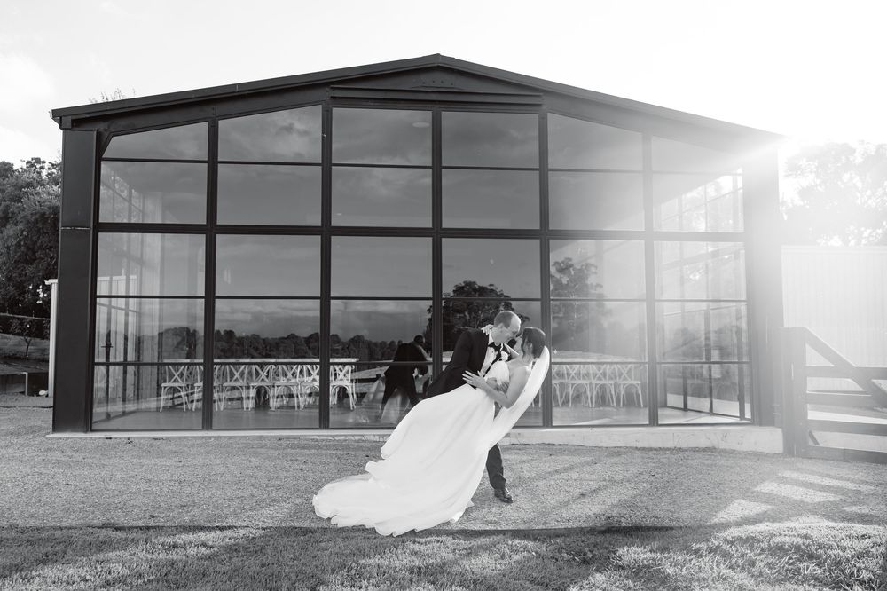 Black and white photo of bride and groom sharing a romantic dip kiss outside a glass pavilion wedding venue.