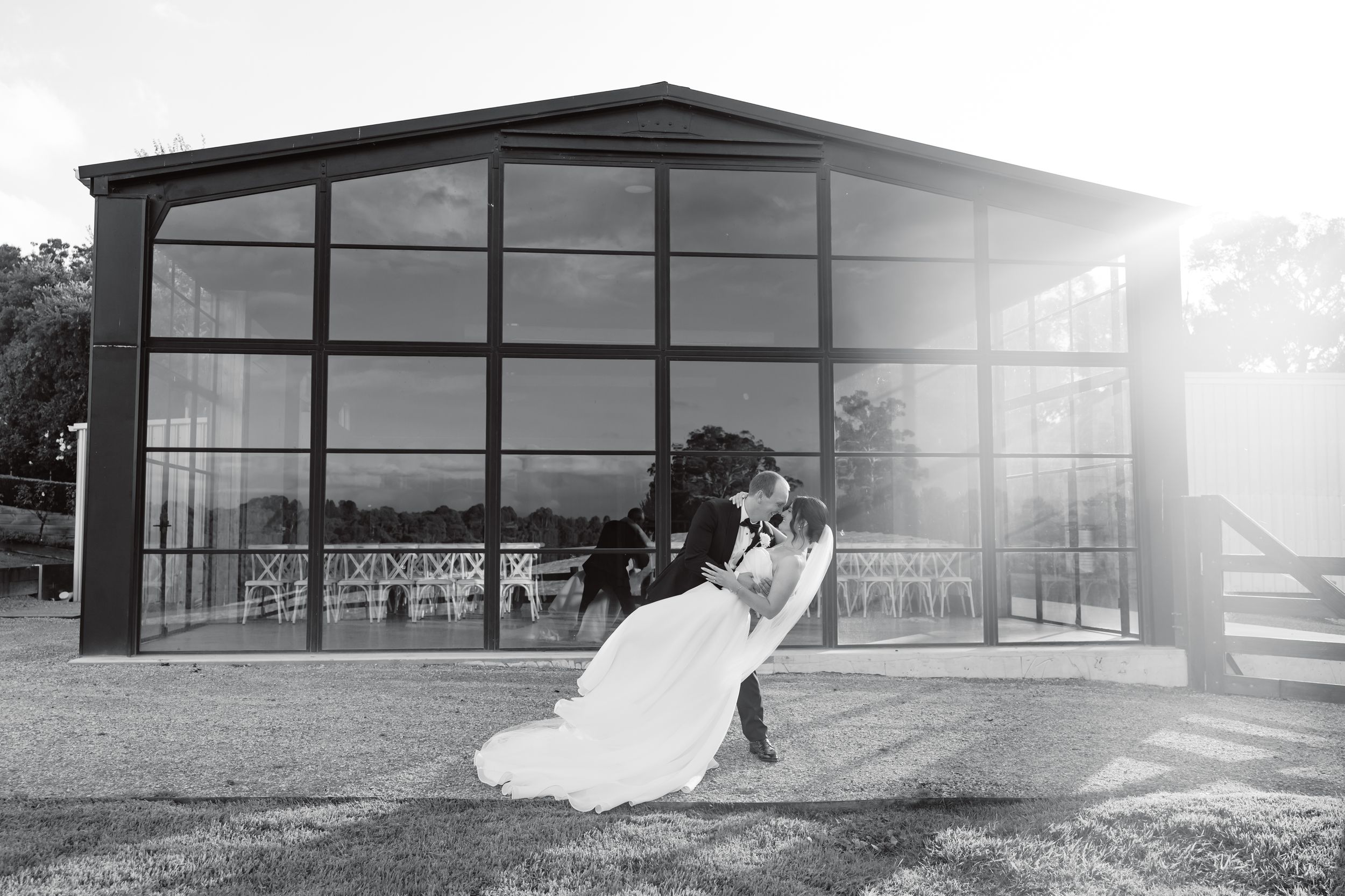 Black and white photo of bride and groom sharing a romantic dip kiss outside a glass pavilion wedding venue.