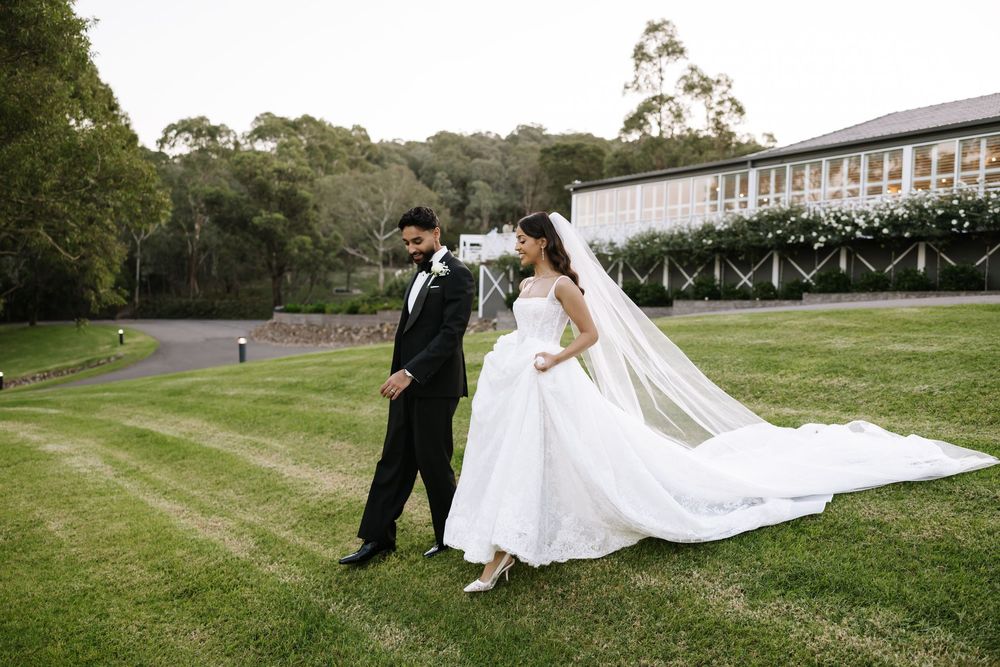 Bride and groom walking on lush green lawn in elegant wedding attire, with a long bridal veil trailing behind.
