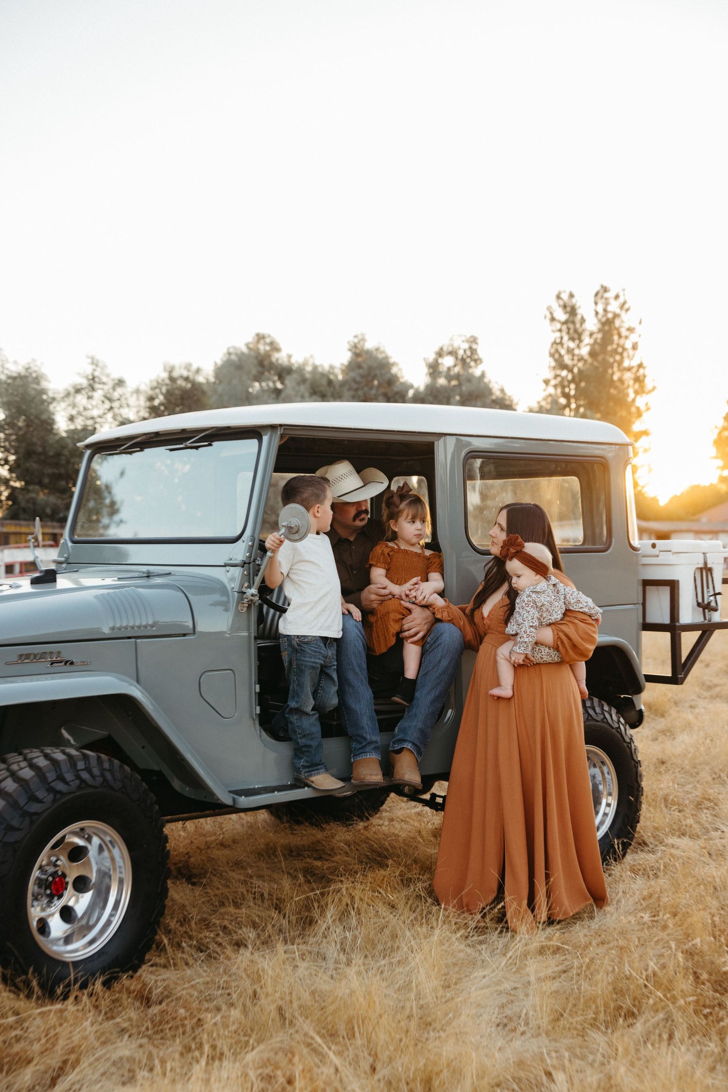 Family gathered around vintage blue Land Cruiser in golden sunset field.