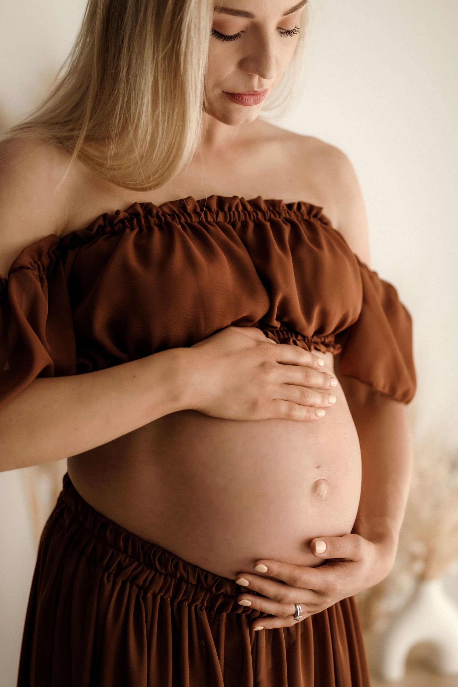 Intimate maternity photo of pregnant woman in white lace lingerie sitting by window with pampas grass.