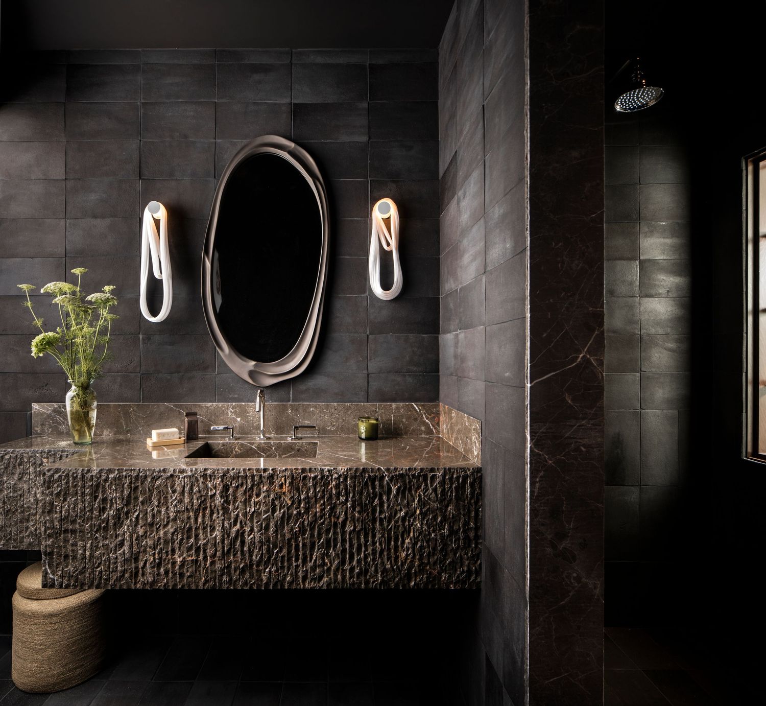 Luxurious dark bathroom with textured stone vanity, oval mirror, modern wall sconces, and sleek walk-in shower.