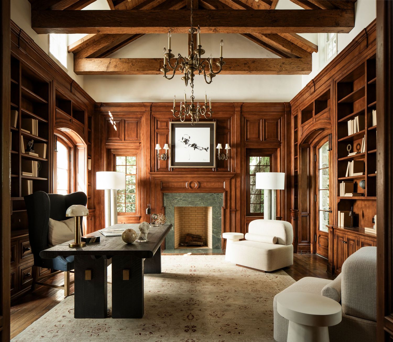 Elegant wood-paneled home library with arched bookshelves, exposed beam ceiling, chandelier, marble fireplace, and modern white chairs.