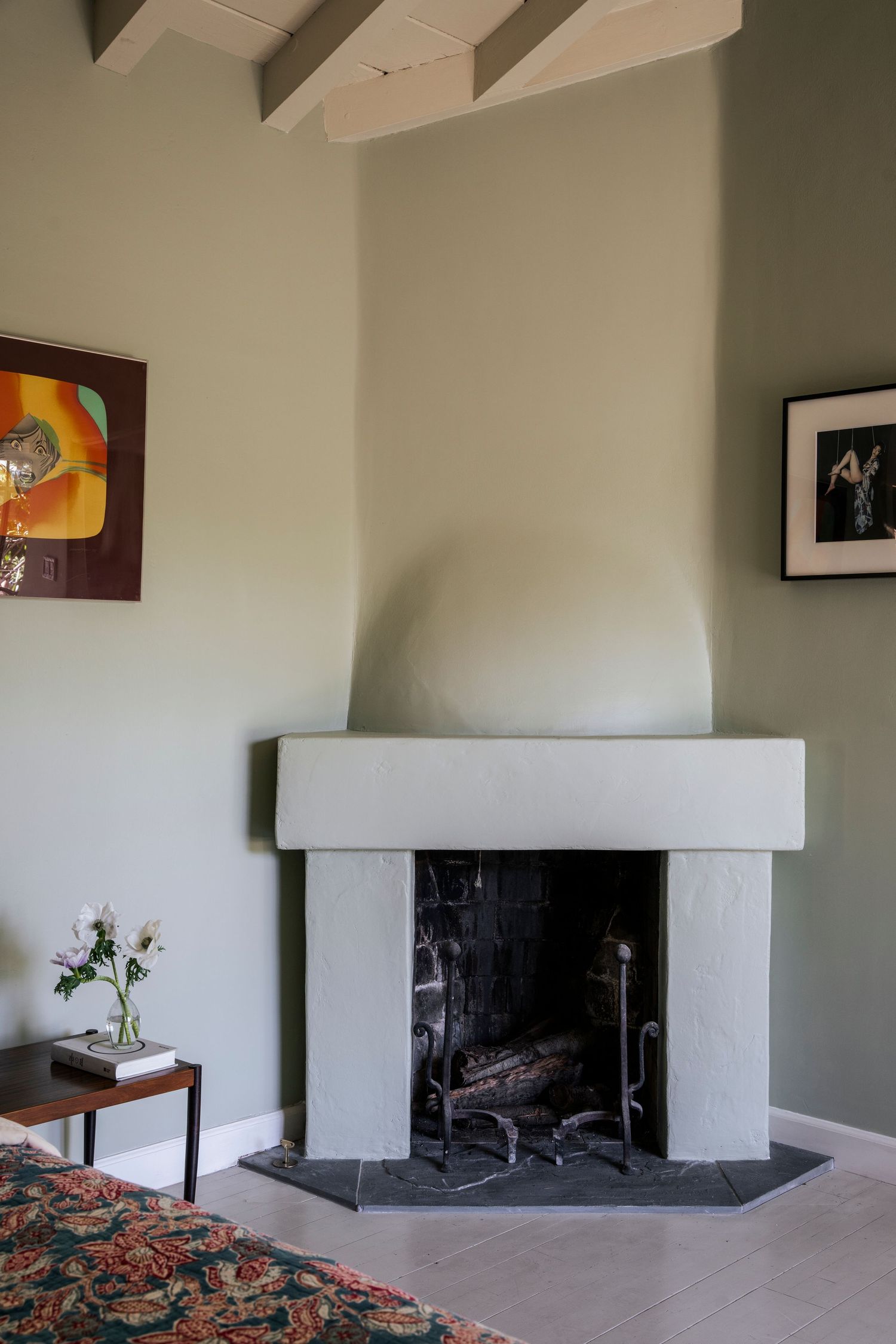 Corner fireplace with sage green walls, exposed beam ceiling, ornate fireplace tools, and colorful artwork in a stylish living room.
