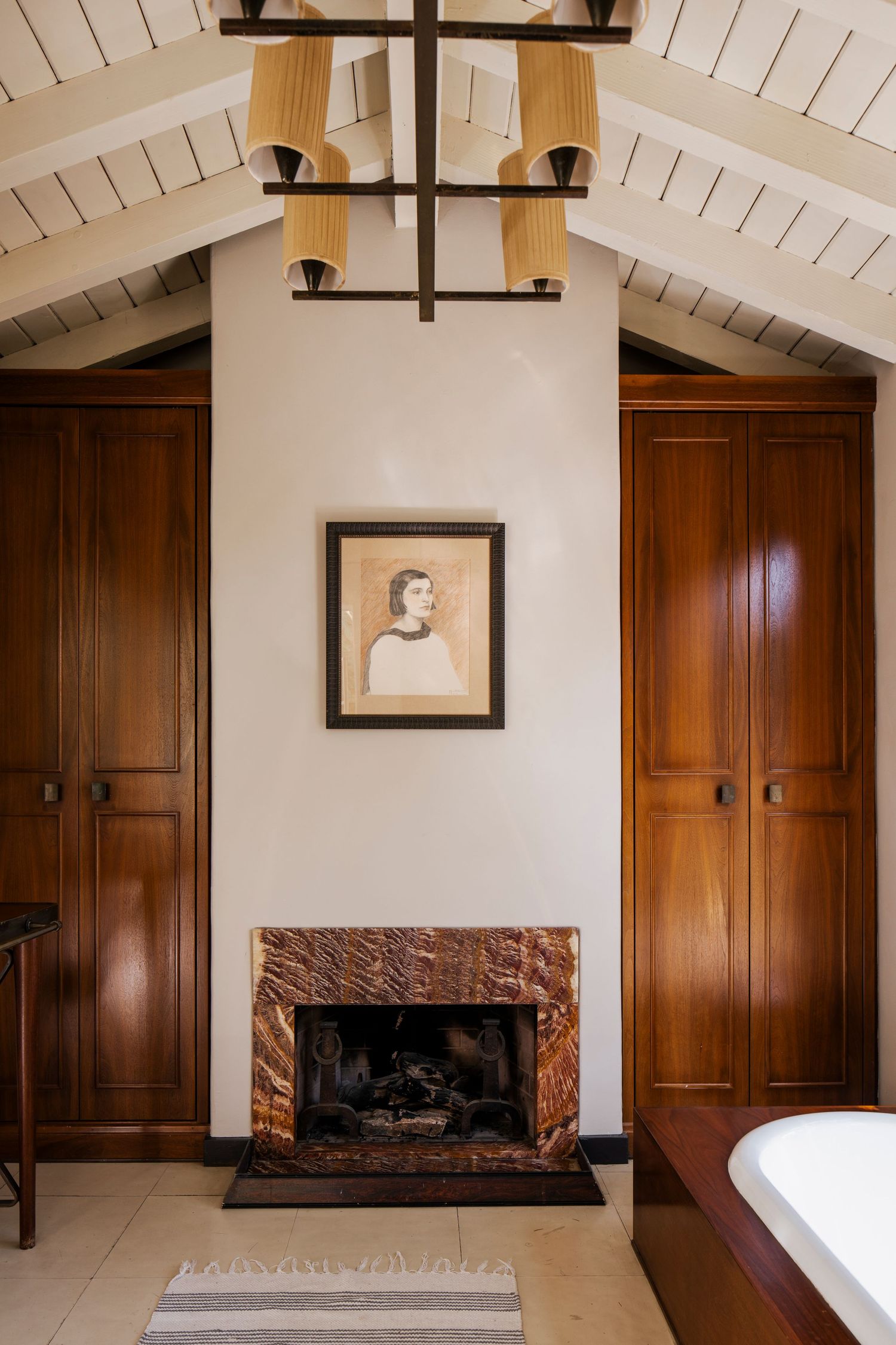 Elegant bedroom with marble fireplace, dark wood wardrobes, vaulted ceiling, chandelier, and portrait artwork above mantel.