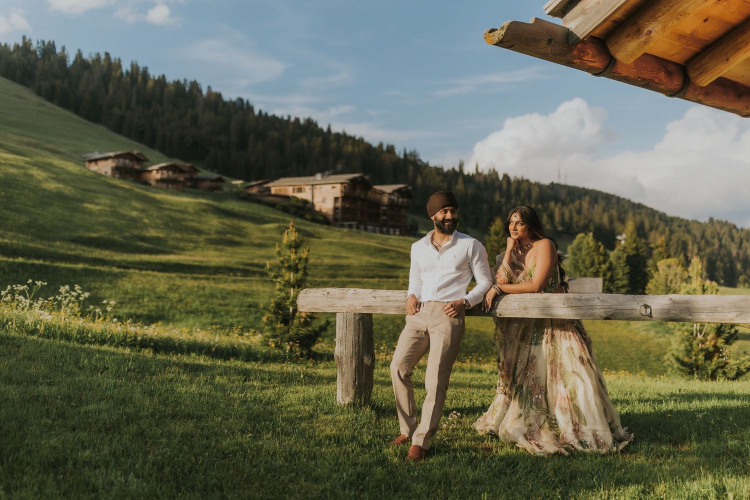 A couple sits on a rustic wooden bench overlooking green mountain meadows and alpine scenery on a sunny summer day.
