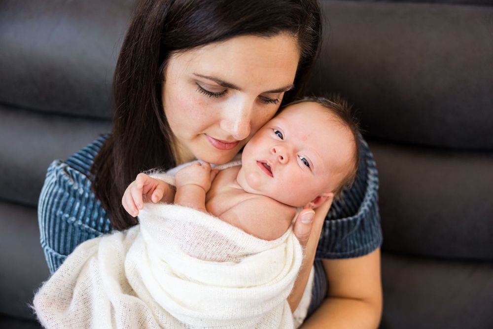 A tender moment between mother and baby wrapped in a soft white blanket on a dark couch.