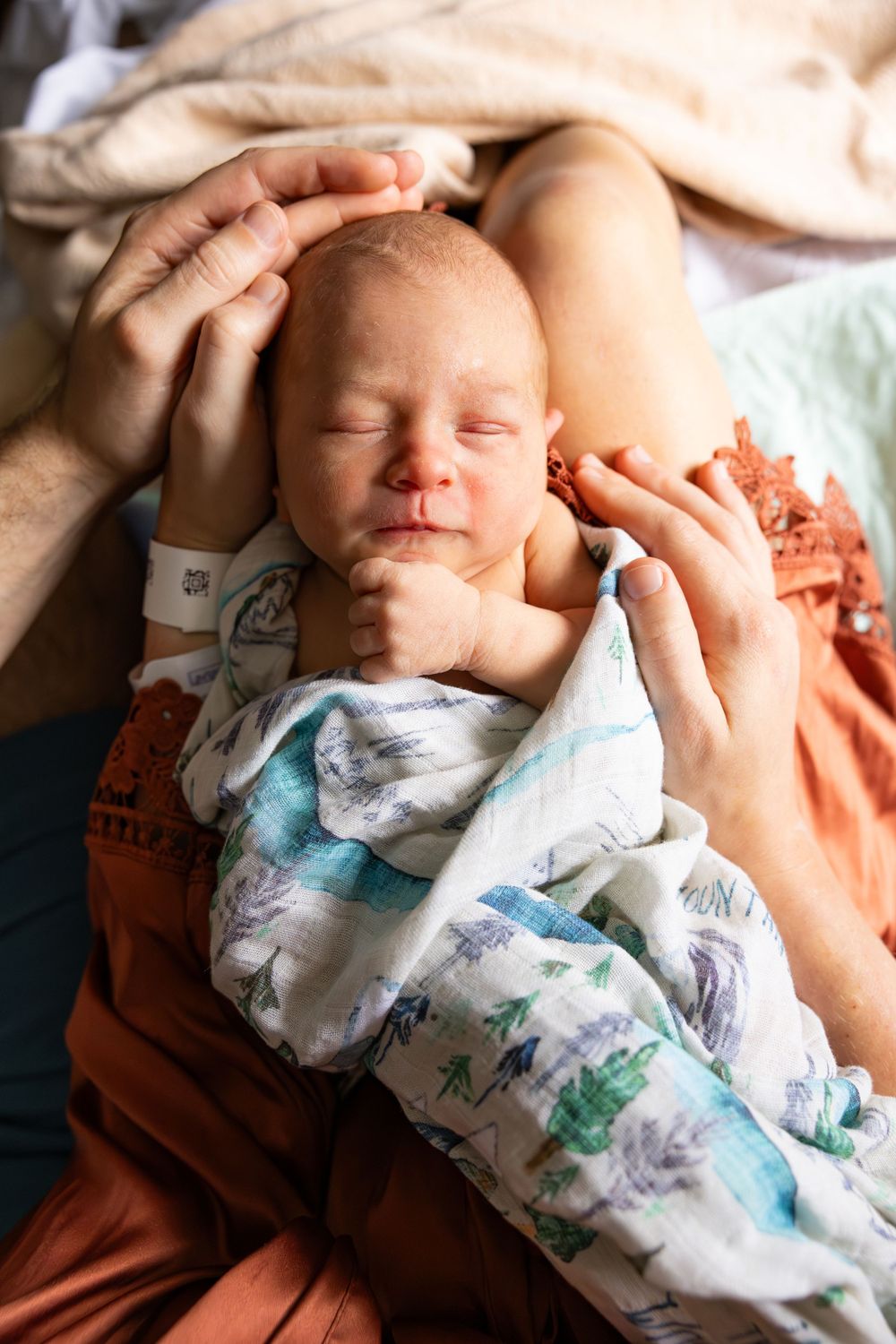 A sleeping newborn baby wrapped in a patterned swaddle blanket while hands tenderly touch their head.