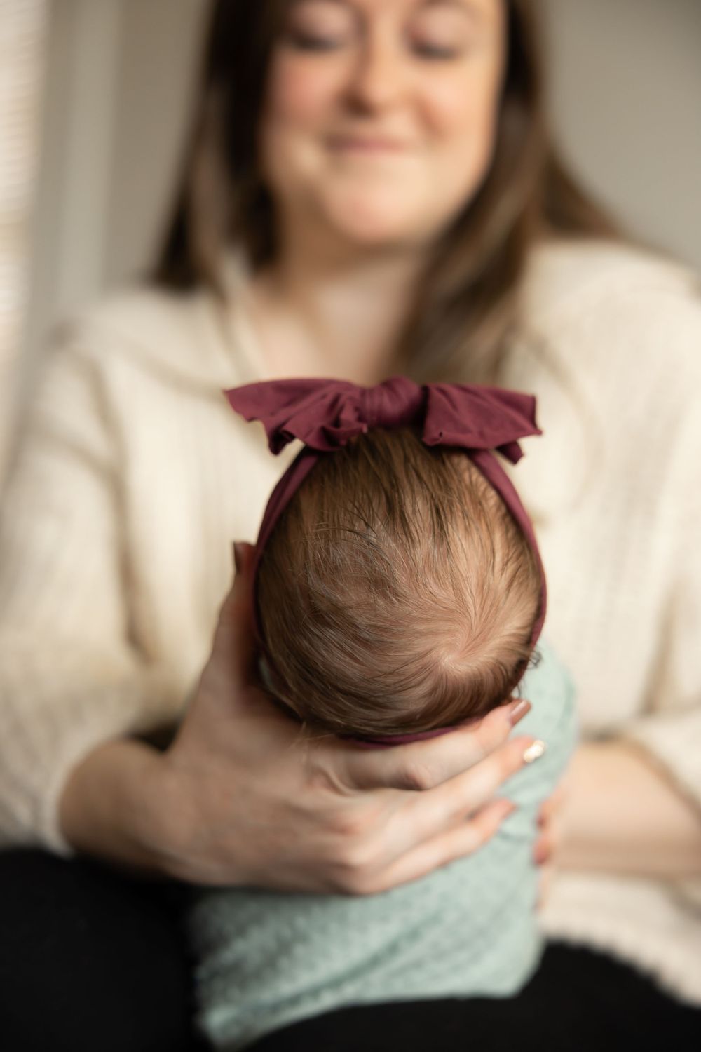 A baby wearing a burgundy bow and mint wrap is held close in a tender moment with a white sweater-clad caregiver.