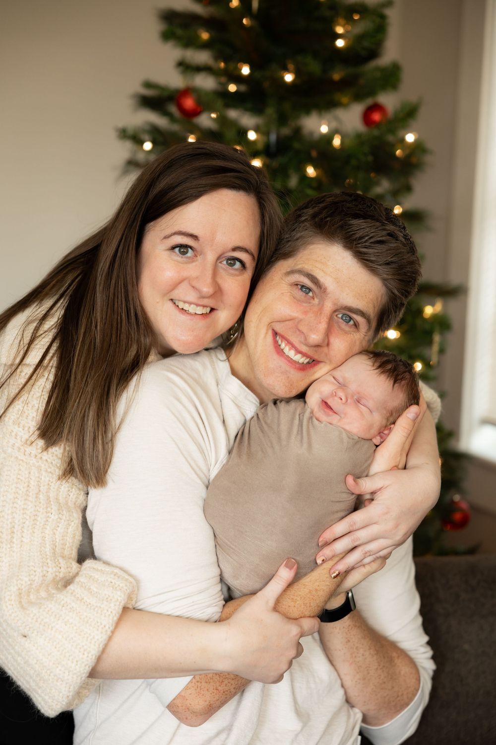A couple embraces and smiles in front of a decorated Christmas tree with lights and red ornaments.