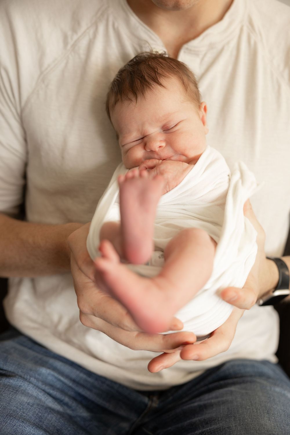 A newborn baby wrapped in white cloth stretches and yawns while being held in someone's lap wearing jeans and white shirt.
