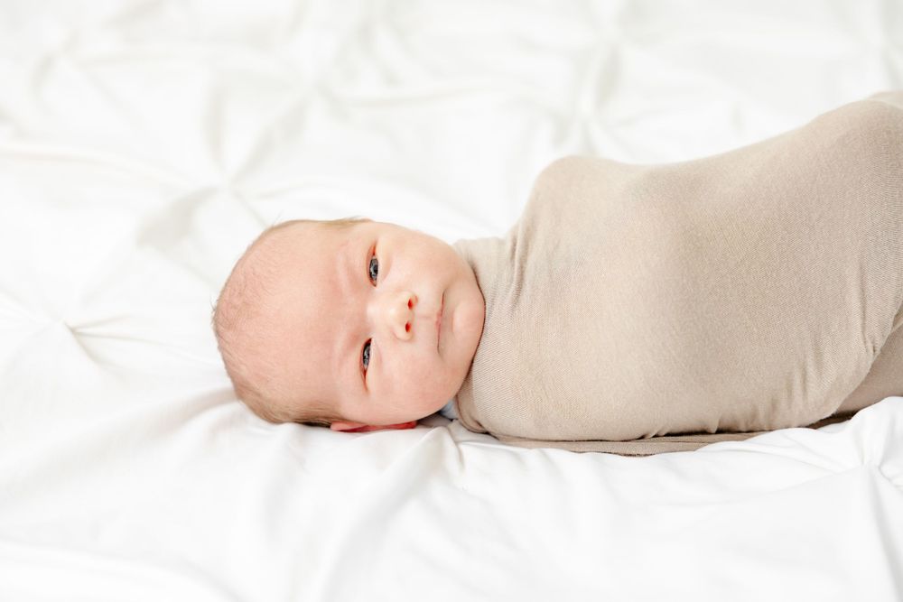 Baby wrapped in beige swaddle blanket lying on white bedding with big eyes looking at camera.