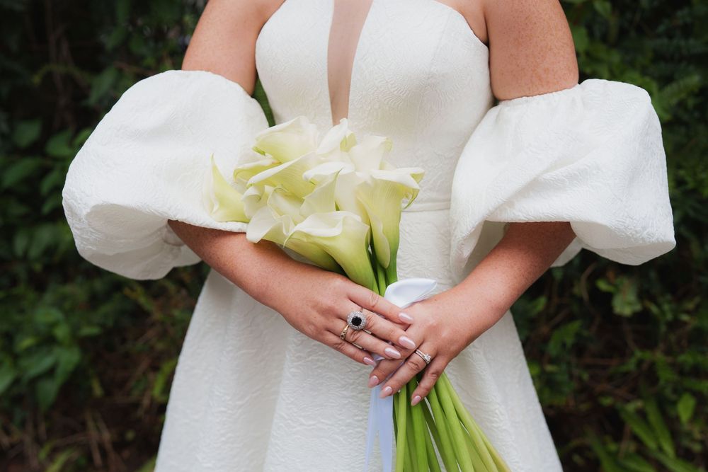 Bride in white gown with puff sleeves holding white calla lily bouquet, wearing sapphire ring.