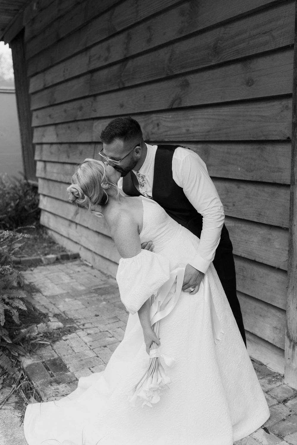 Black and white photo of a groom dipping and kissing his bride in a white gown outside a wooden barn.