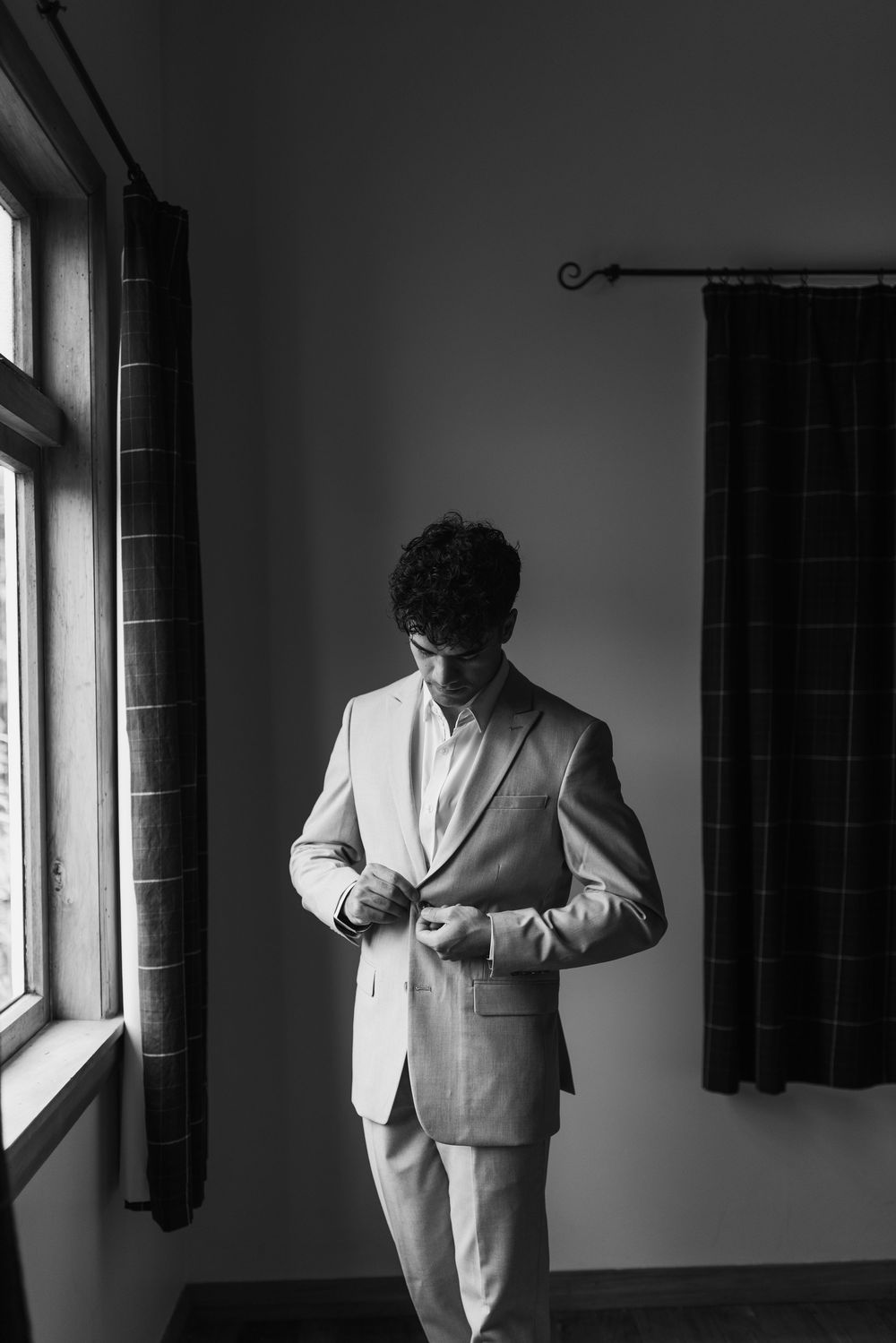 Black and white photo of a groom buttoning his suit jacket while standing near a window before his wedding.